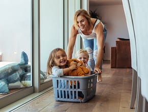 Happy young mother pushing children sitting in laundry basket. Mother and children playing at home.