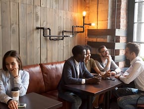 Diverse young friends ignoring sad girl sitting alone in cafe